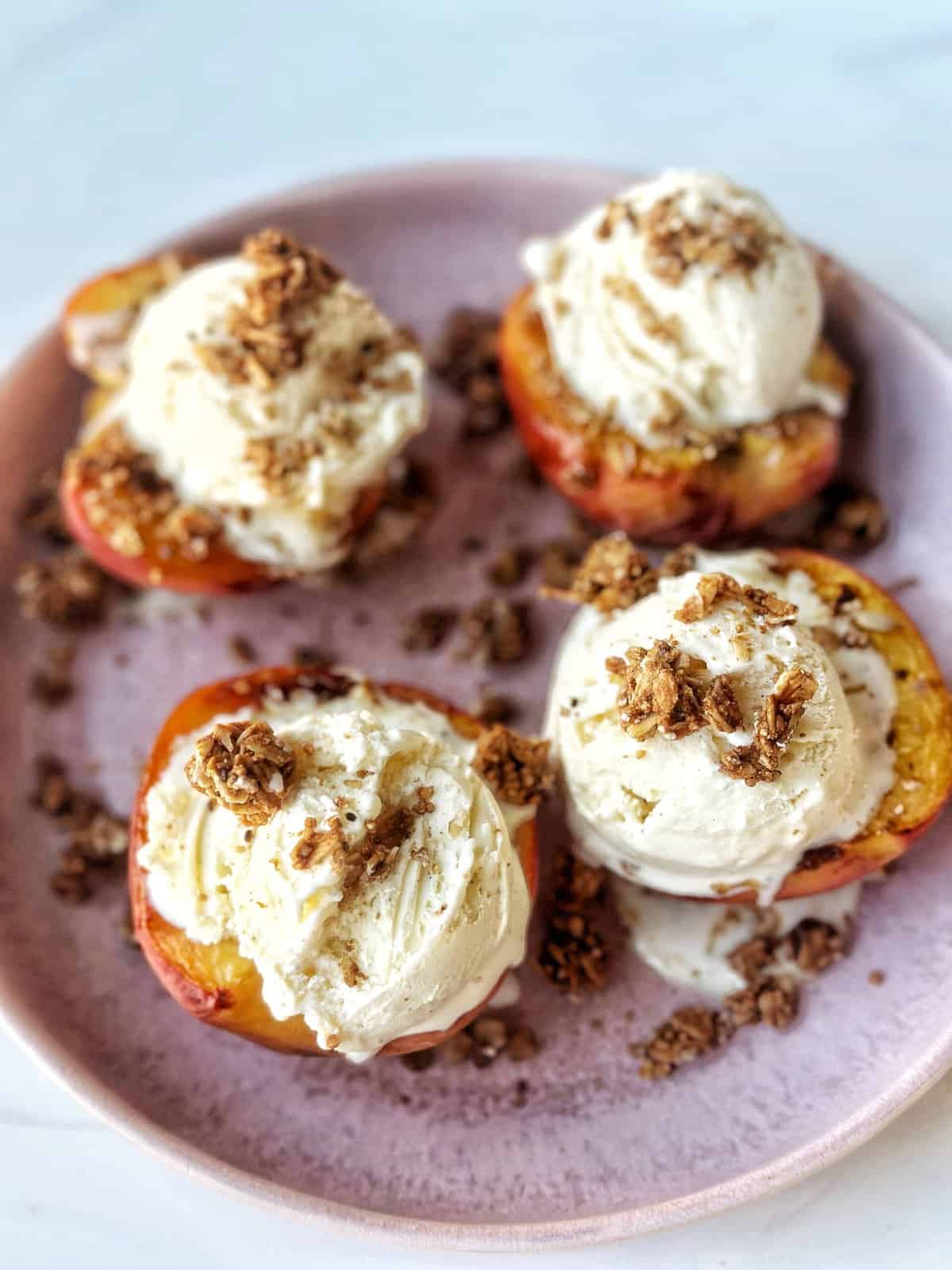 Juicy peach halves with vanilla ice cream and granola topping on pink plate.