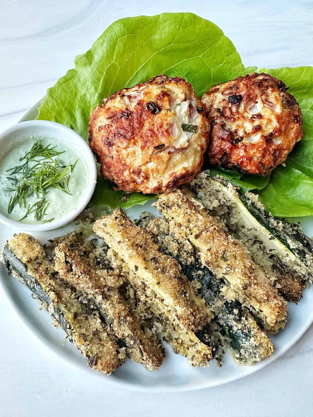 Crunchy breaded zucchini fries with homemade garlic sauce and savory chicken burgers on a white plate.