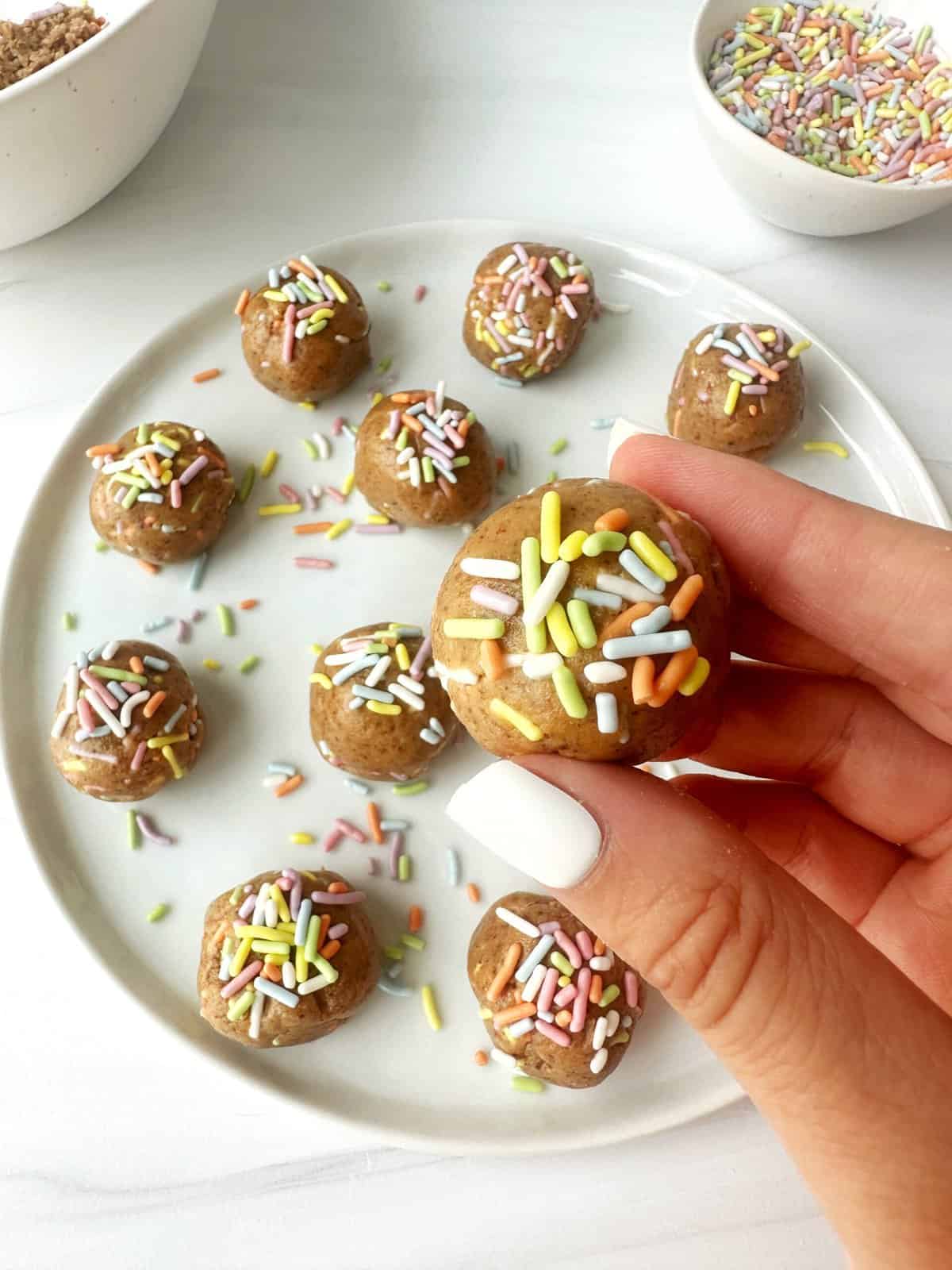 Colorful sprinkle-topped edible cookie dough bites on white plate.