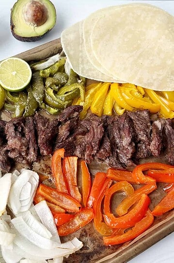 Juicy carne asada with colorful grilled peppers, sliced avocado, and fresh lime on a rustic platter.
