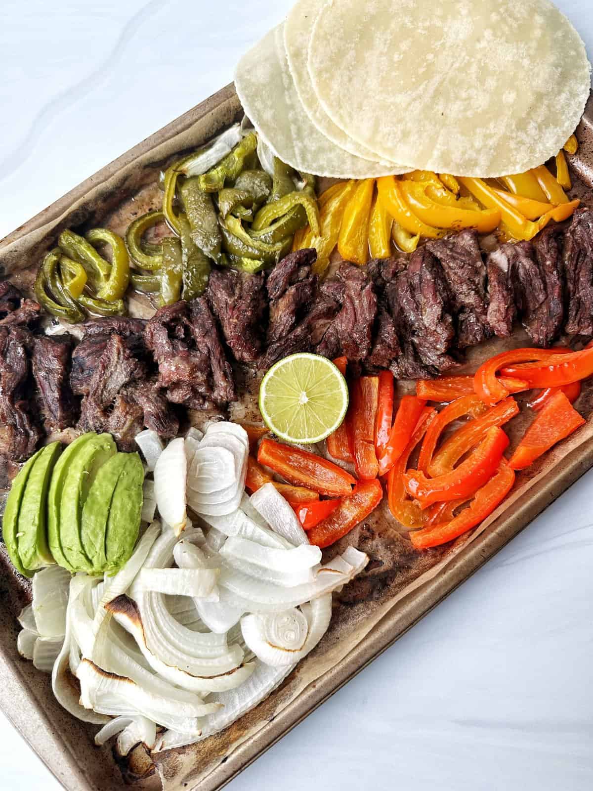 Succulent grilled steak with colorful peppers, onions, avocado, and fresh lime on a sheet pan