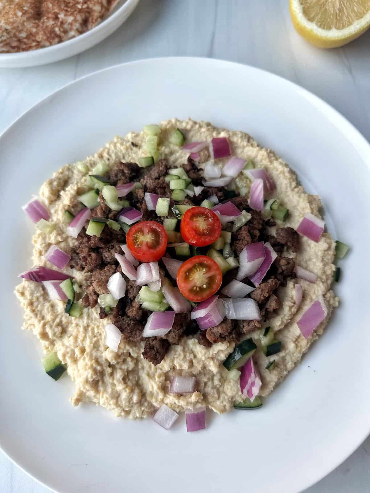 Creamy hummus topped with cooked ground beef, chopped cucumbers, cherry tomatoes, and red onions on a white plate.