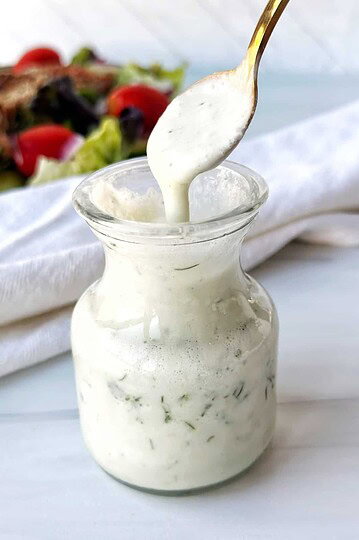 Creamy homemade ranch dressing in a glass jar on a white surface.
