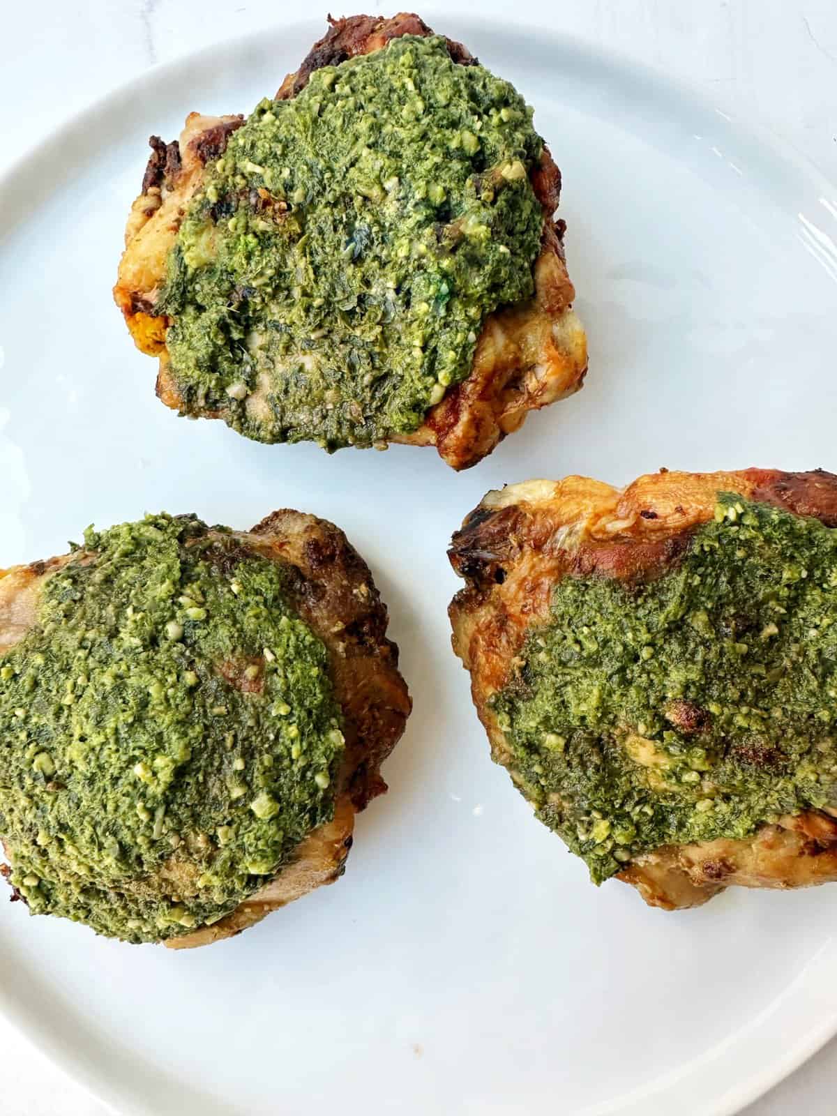 Tender baked chicken thighs topped with vibrant basil pesto, served on a white plate.