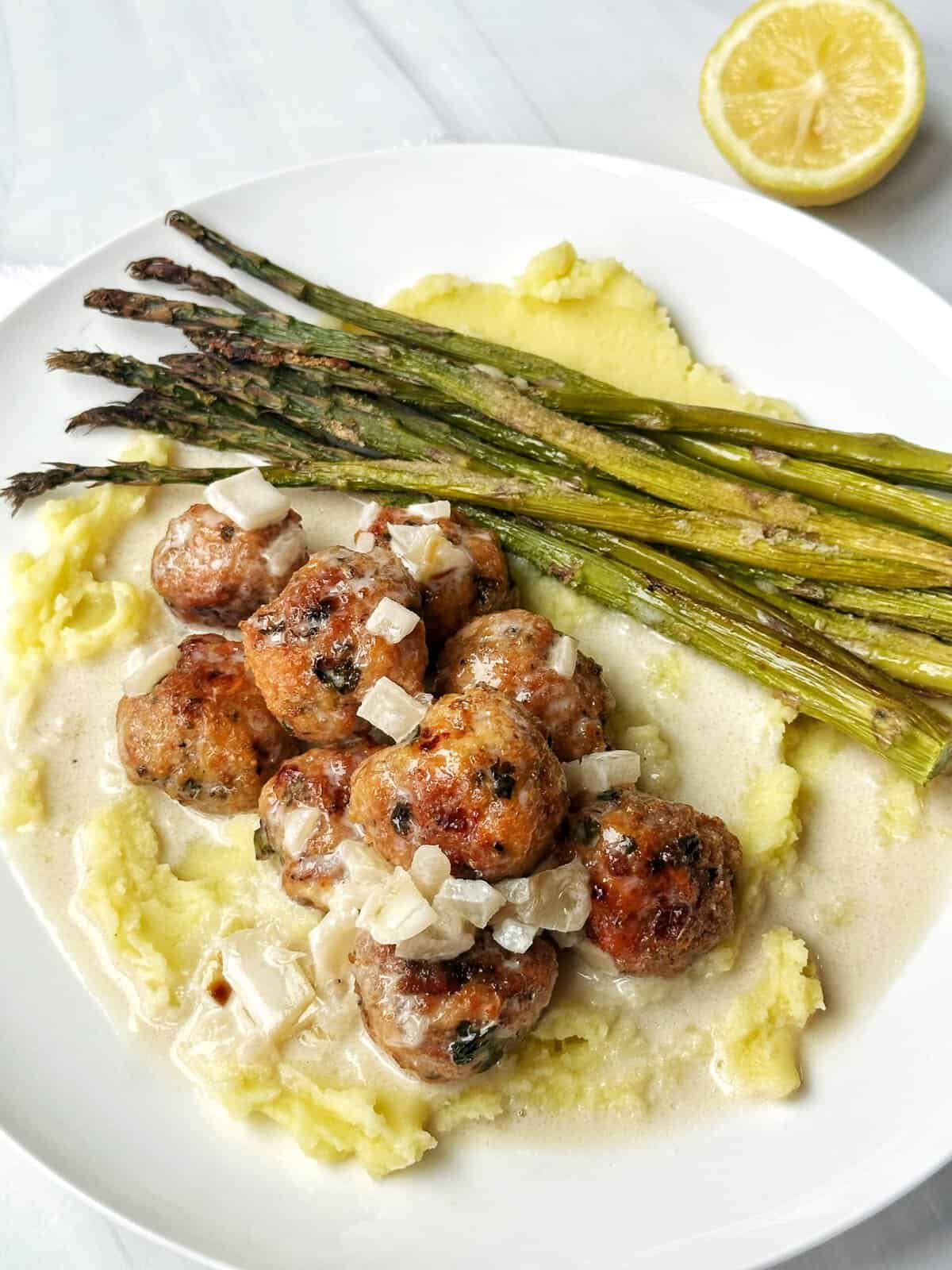 Roasted asparagus, creamy mashed potatoes, and Swedish meatballs with white sauce and onions on a white plate.
