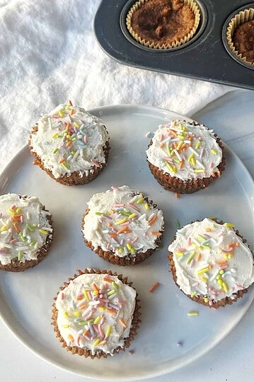 Colorful frosted cupcakes with sprinkles on white plate, with baking tray in background.