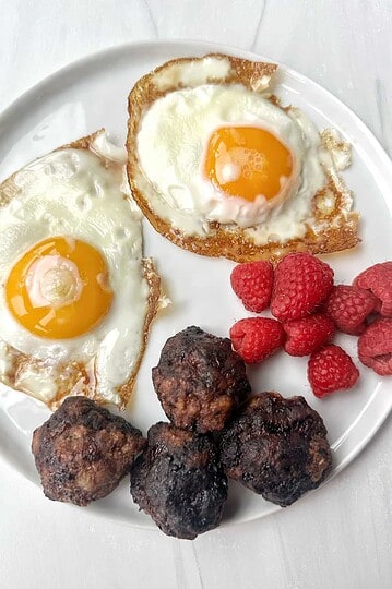 Eggs sunny side up with homemade meatballs and fresh raspberries on a white plate.