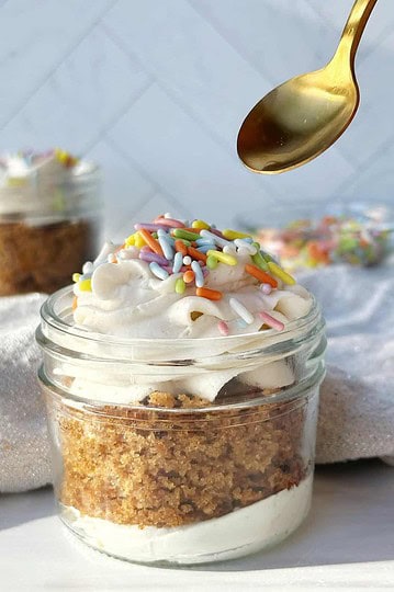 Cream cheese frosting with colorful sprinkles layered over classic cheesecake crust in a glass jar.