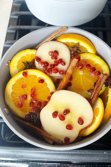 Fresh sliced oranges and apples with cinnamon sticks and pomegranate seeds in a bowl, perfect for cozy winter beverages.