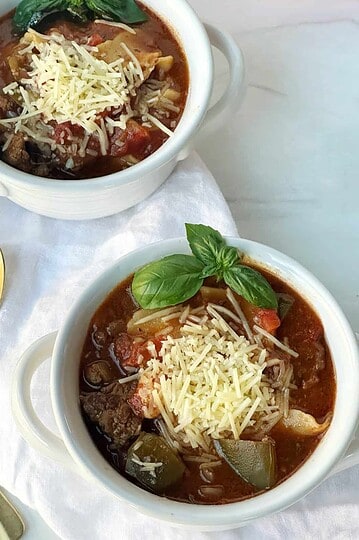 Beef and vegetable soup in white ramekins with shredded cheese and fresh basil.