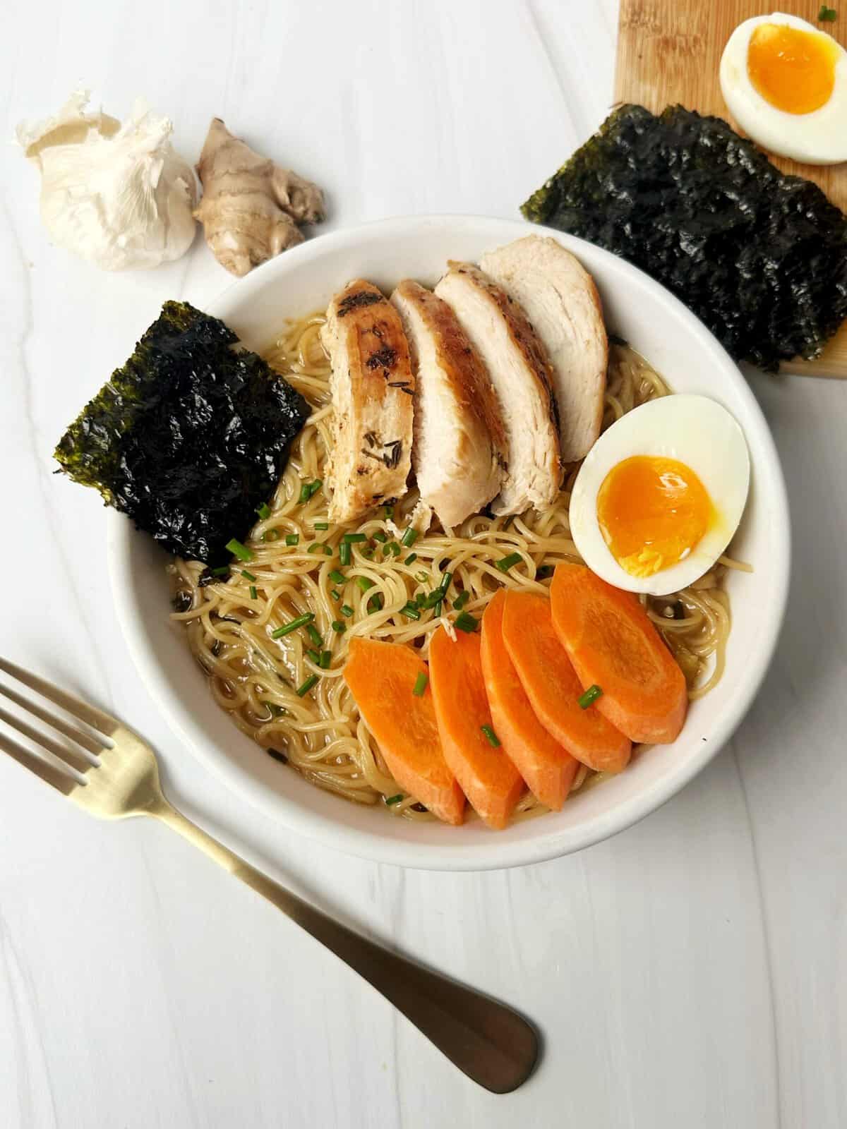 Creamy ramen bowl with grilled chicken, soft-boiled eggs, seaweed, and fresh vegetables on white table.