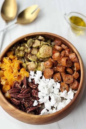 Savory meal bowl with roasted sweet potatoes, sautéed brussels sprouts, vegan sausage, pecans, and crumbled feta cheese.