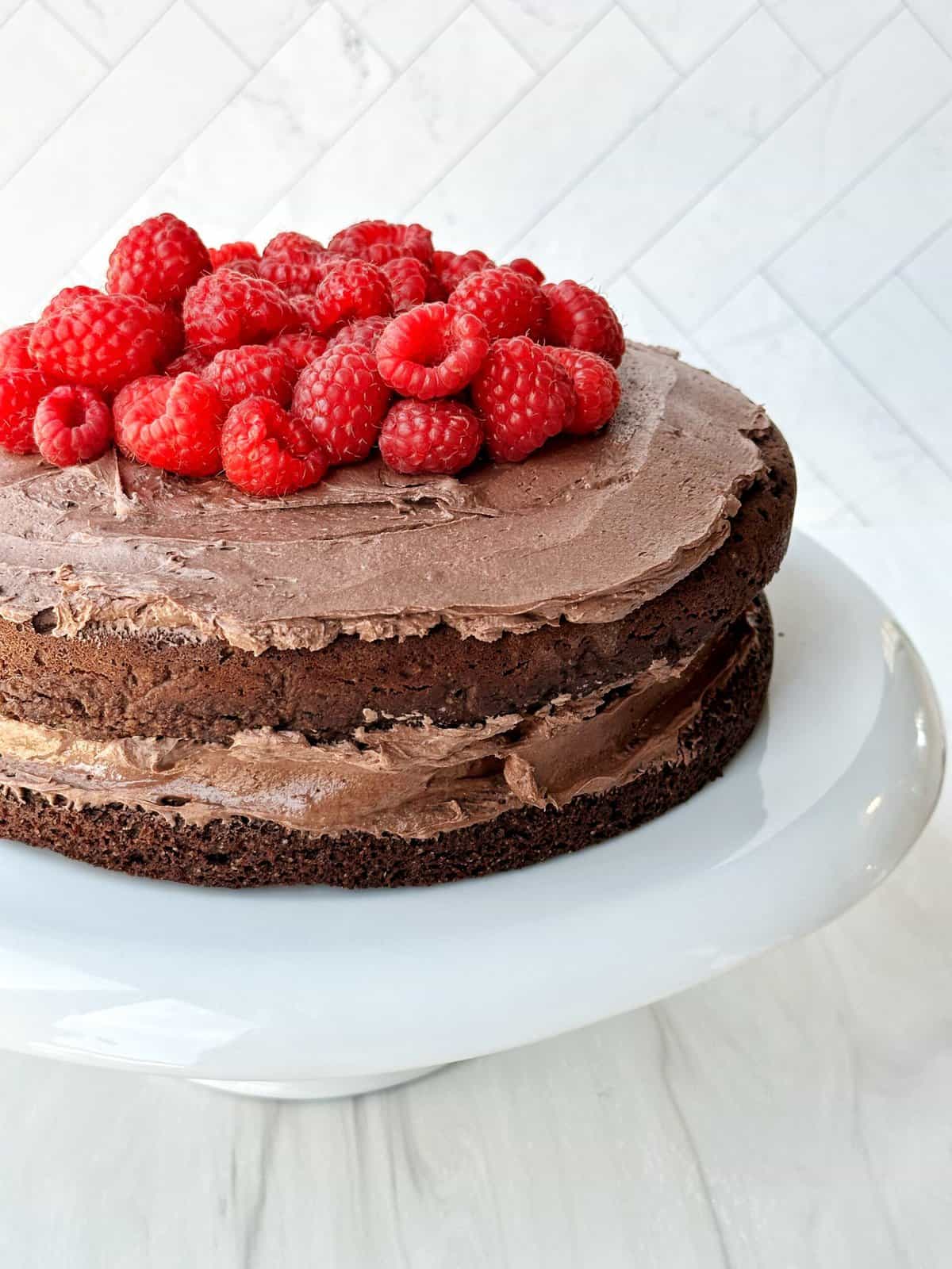 Fresh chocolate cake topped with raspberries on a white cake stand.