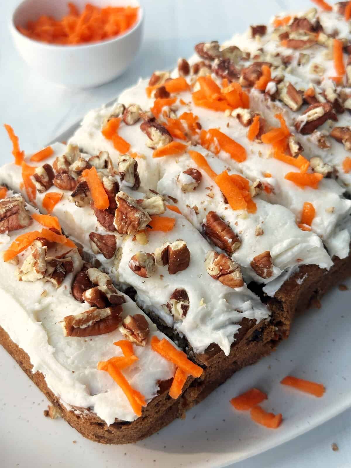 Cream cheese carrot cake with chopped pecans and shredded carrots on top, served on a white platter.