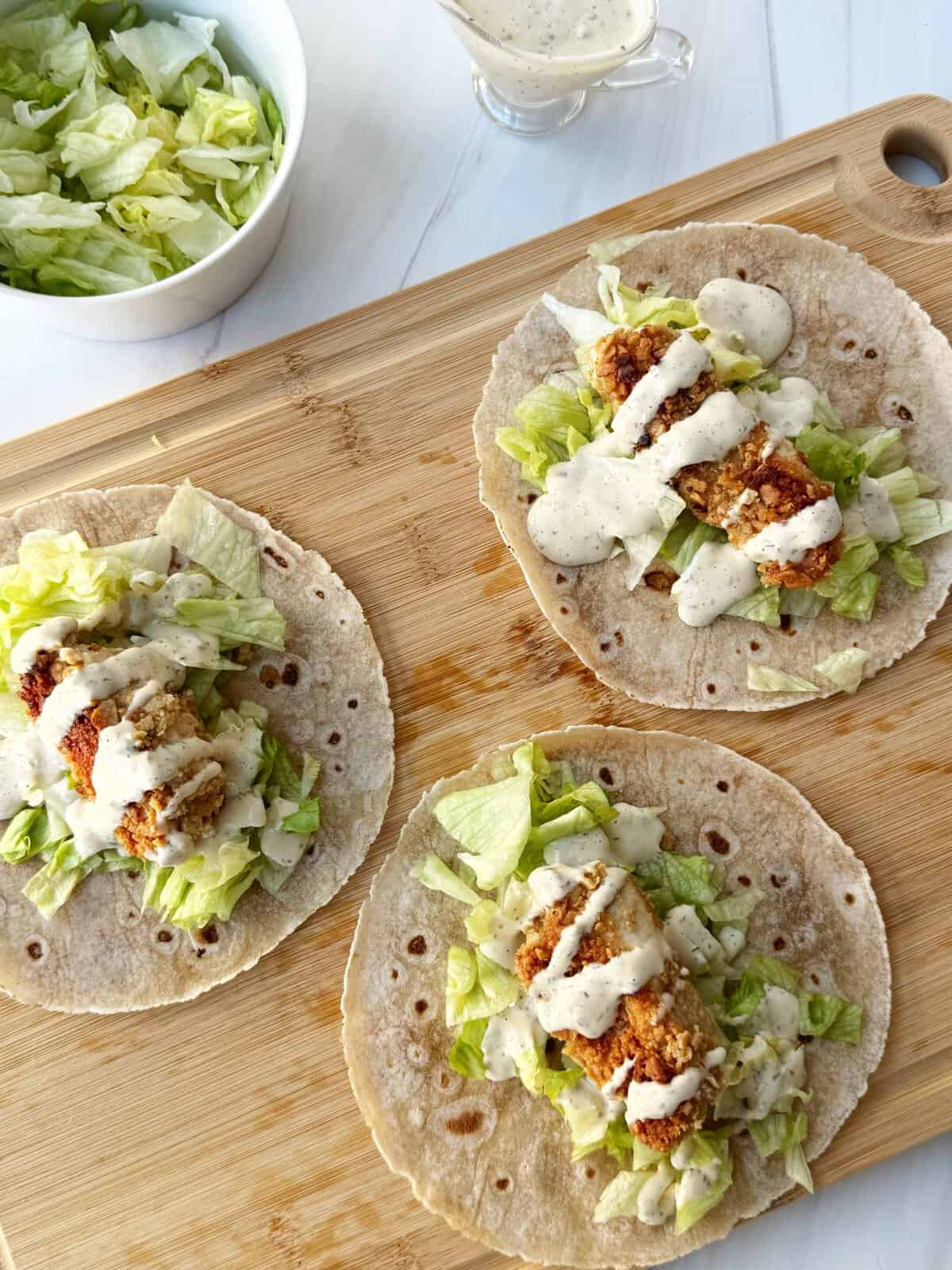 Fried chicken tacos with lettuce and creamy sauce on wooden cutting board.