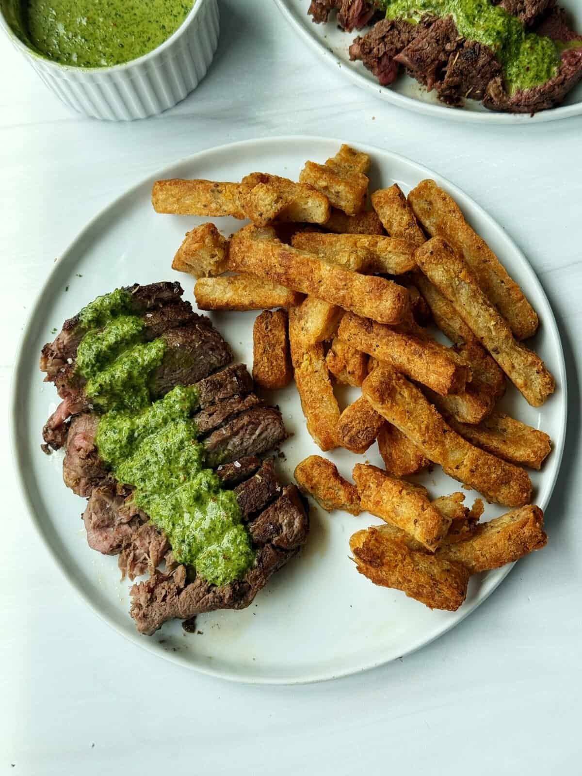 Juicy steak with vibrant green herb sauce and crispy French fries on a white plate.