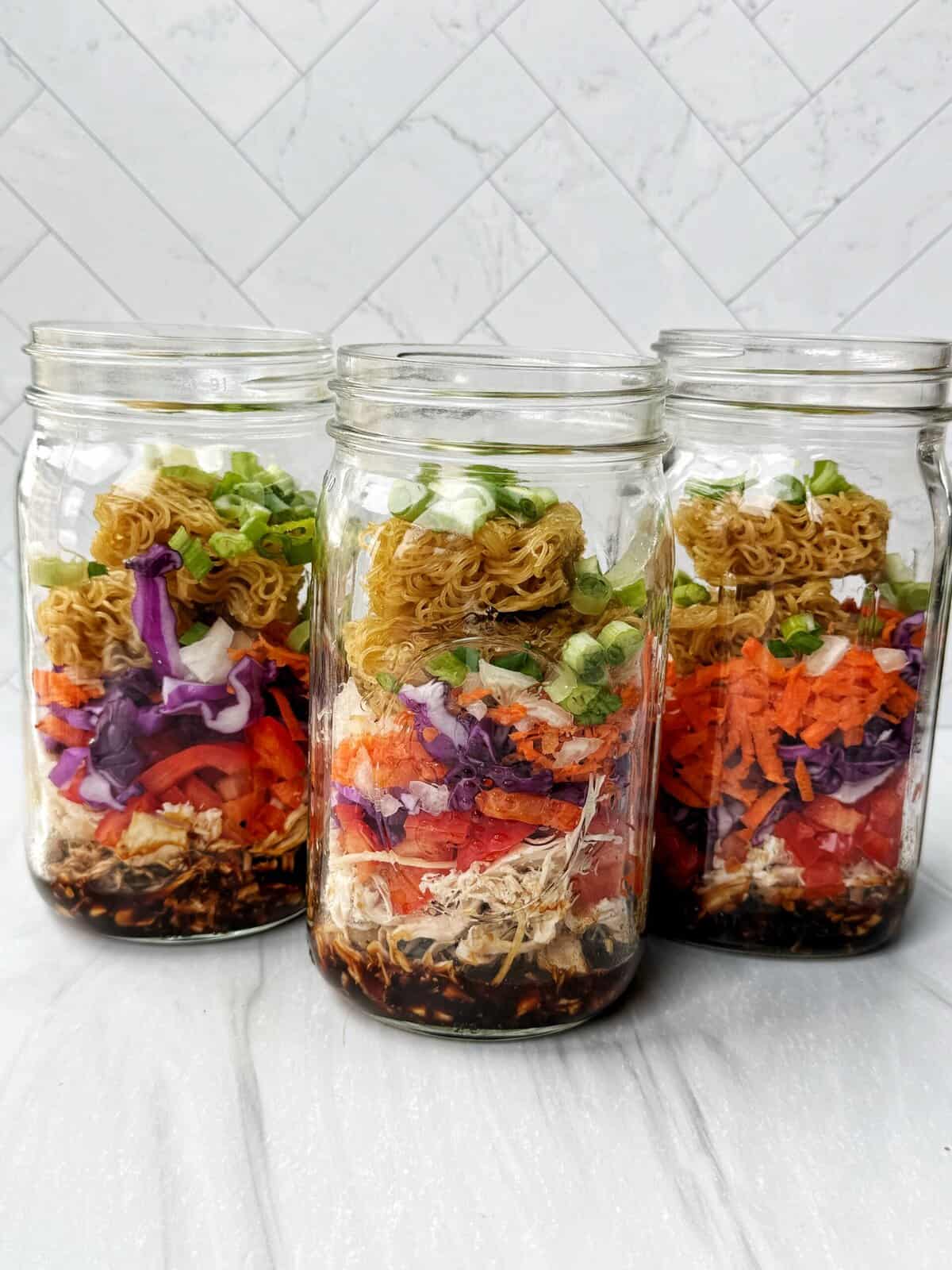 3 glass jars with sauce, chicken, vegetables and ramen noodles layered