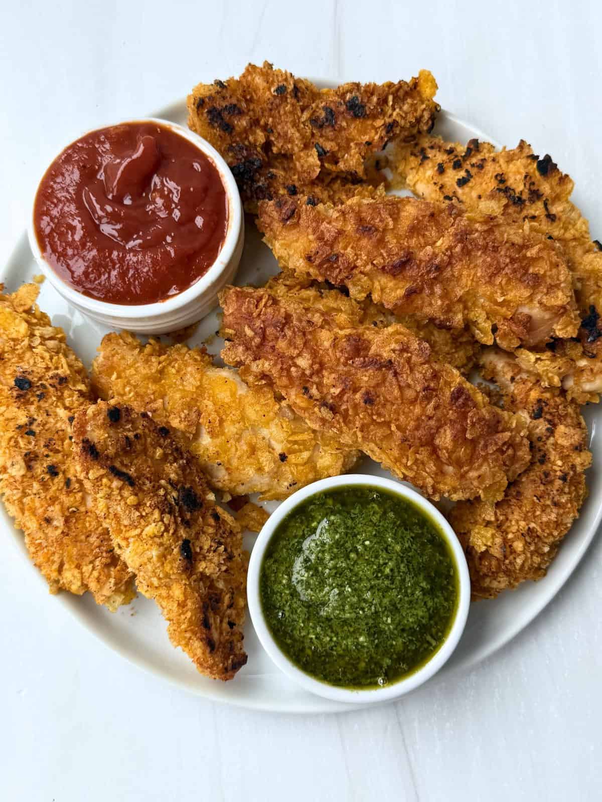 crispy chicken tenders on a white plate with ketchup and pesto dipping sauces 